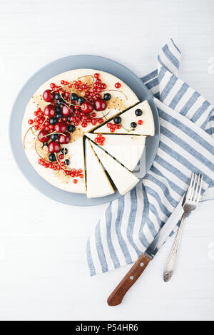 Hausgemachter Käsekuchen mit Beeren auf weiße Holztisch mit Handtuch, Messer und Gabel. Ansicht von oben. Rote Johannisbeere, schwarze Johannisbeere und Kirsche. Stockfoto