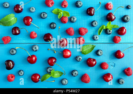 Beeren auf blauem Hintergrund, flach. Berry Muster auf blauem Holz Hintergrund. Himbeere, Kirsche, Heidelbeere und Mint Blatt Muster Stockfoto