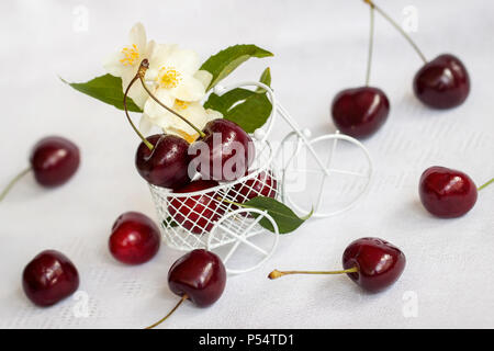 Kleines Spielzeug Warenkorb, Trolley Nahaufnahme reife Kirsche, Jasmin Blumen auf hellem Hintergrund. Vegetarische Konzept, Ernährung, Entgiftung, organische Vitamine Stockfoto