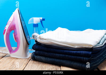 Bügeleisen und Stapel von Jeans und T-Shirt Kleidung auf Holzbrett, blauer Hintergrund Stockfoto