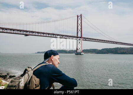 Ein junger Reisender oder ein Tourist mit einem Rucksack auf der Uferpromenade in Lissabon in Portugal am 25. April Brücke Stockfoto