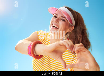 Glückliche junge Frau im gelben Hemd gegen den blauen Himmel, die Herzförmigen Hände Stockfoto