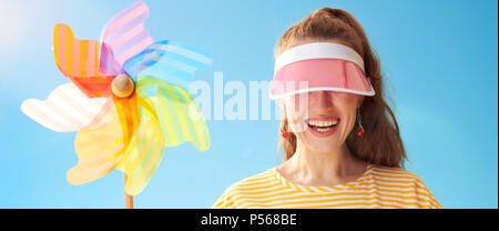 Junge Frau im gelben Hemd gegen den blauen Himmel verstecken hinter der Sonnenblende Holding bunte Mühle Stockfoto