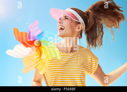 Freundliche gesunde Frau im gelben Hemd gegen den blauen Himmel spielen mit bunten Windmühle Stockfoto