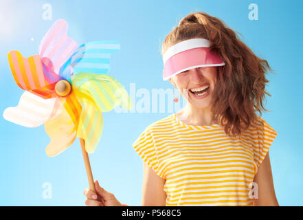 Fröhliche junge Frau im gelben Hemd gegen den blauen Himmel verstecken hinter der Sonnenblende Holding bunte Mühle Stockfoto