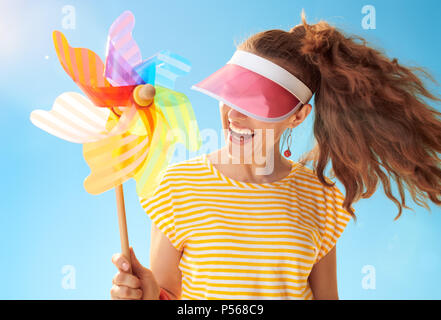 Freundliche gesunde Frau im gelben Hemd gegen den blauen Himmel verstecken hinter der Sonnenblende Holding bunte Mühle Stockfoto