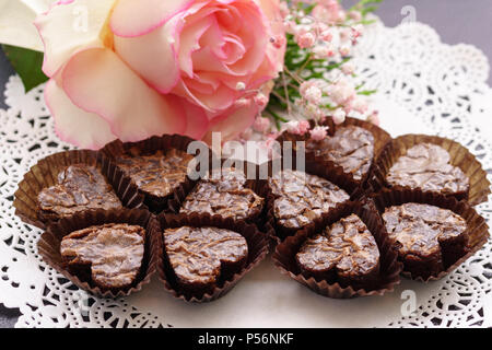 Herzförmige brownies in Pergamyn braun Pappbecher auf einem weißen Deckchen, verziert mit einer weißen Rose mit rosa Grenzen. Stockfoto