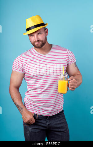 Kreative männlichen im Sommer gestreiftes Hemd mit Cocktail Flasche entspannt und Stroh Hut gelb auf blau studio Hintergrund Stockfoto