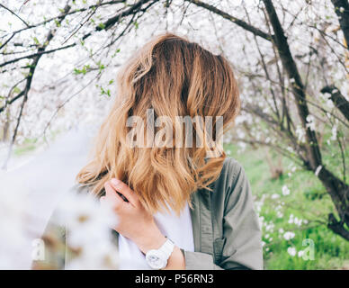Junge Frau mit Flatternden blondes Haar steht neben einem blühenden Bäume. Candid lifestyle Foto des modischen weiblichen Frühlingstag. Stockfoto