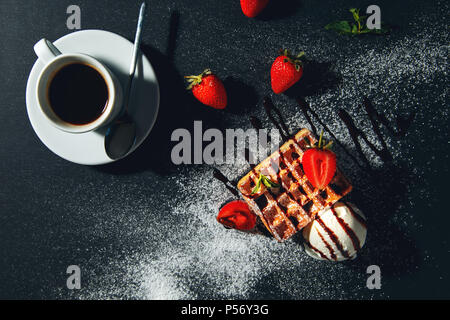 Tasse Kaffee mit belgischen Waffeln mit Eis und Erdbeeren auf einem schwarzen Hintergrund. Stockfoto