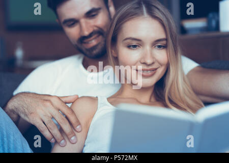 Die schöne Zeit zu Hause. Schöne junge Liebende Paar kleben aneinander und lächelnd, während Frau mit Buch Stockfoto