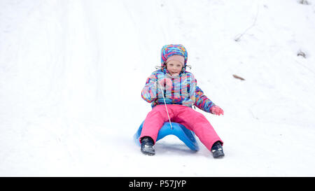 Fröhliches Mädchen reitet einen Schlitten bergab auf einer Schnee bedeckten Schlitten Trail in einer weißen sonnigen Winter Berglandschaft Stockfoto