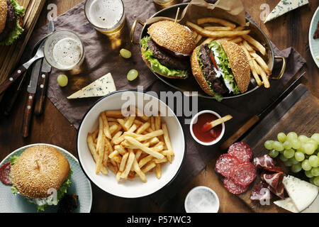 Burger und Pommes frites in Pfannen auf Holztisch mit Soße, Bier, Salat und verschiedene Snacks top Aussicht serviert. Tisch Konzept Stockfoto