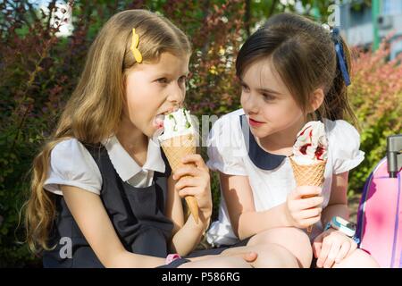 Porträt von zwei Freundinnen Schülerinnen im Alter von 7 Jahren in der Schule Uniform mit Rucksäcken Eis essen. Hintergrund Stadt, Sommer, Gehweg, dekorative b Stockfoto