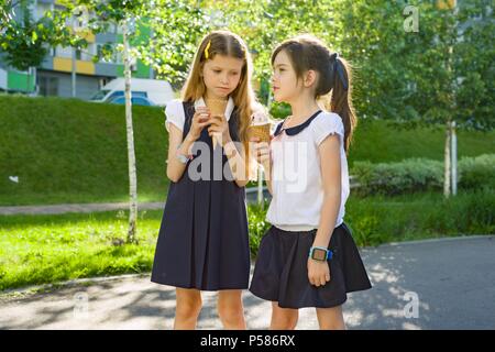 Porträt von zwei Freundinnen Schülerinnen im Alter von 7 Jahren in Schuluniform Eis essen. Hintergrund Stadt, Sommer, Gehweg, dekorative Sträucher. Stockfoto