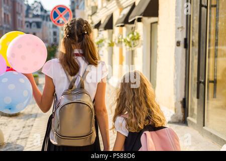 Zwei Mädchen sind zu Fuß entlang der Straße mit bunten Luftballons. Kinder in Schuluniform, Gläser, Rucksack. Ansicht von hinten. Stockfoto