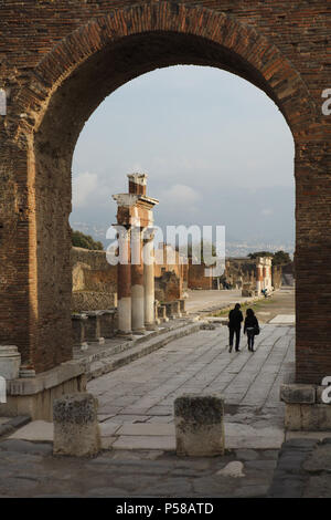 Paar Besucher gehen auf das römische Forum in die archäologische Stätte von Pompeji (Pompei) in der Nähe von Neapel, Kampanien, Italien, dargestellt durch das Römische Triumphbogen. Stockfoto