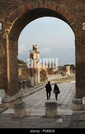 Paar Besucher gehen auf das römische Forum in die archäologische Stätte von Pompeji (Pompei) in der Nähe von Neapel, Kampanien, Italien, dargestellt durch das Römische Triumphbogen. Stockfoto