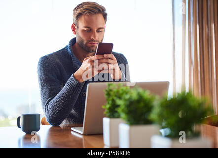 Hübscher junger Mann eine SMS auf seinem Mobiltelefon während an seinem Esstisch in der von seiner Wohnung eine frische Tasse Kaffee und einen modernen Laptop auf dem Tisch vor ihm sitzt. Stockfoto