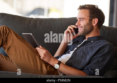 Close up Profil Image der jungen attraktiven kaukasischen Mann lächelnd, während Sie telefonieren und halten eine Tablette in seiner anderen Hand, während Sie mit einem großen Mode Sinn und eine stilvolle Uhr gekleidet. Stockfoto