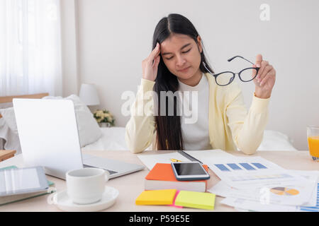 Asiatische Frau Freiberufler Kopfschmerzen und Stress während der Arbeit mit dem Laptop auf dem Tisch im Schlafzimmer zu Hause. Arbeit zu Hause Konzept. Arbeiten von zu Hause Konzept Stockfoto