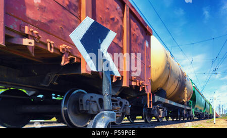 Ein Güterzug mit Güterwagen und Zisternen eilt hinter dem Pfeil gegen den blauen Himmel im Sommer Stockfoto