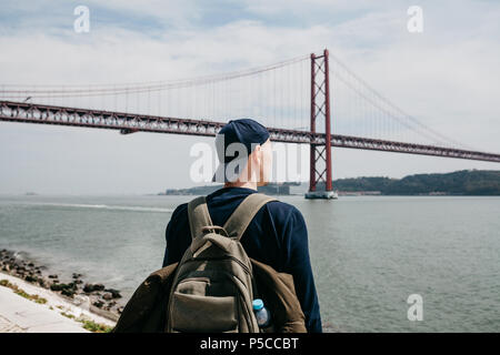 Ein junger Reisender oder ein Tourist mit einem Rucksack auf der Uferpromenade in Lissabon in Portugal am 25. April Brücke Stockfoto