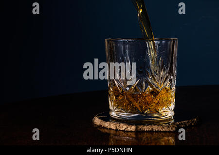 Whiskey in einem Kristall Tumbler auf einem dunklen Holztisch mit einem blauen Hintergrund gegossen. Mit Kopie Raum auf der linken Seite. Stockfoto