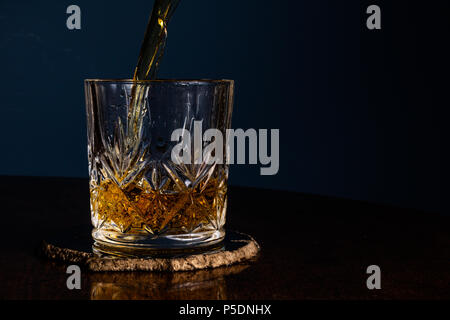 Whiskey in einem Kristall Tumbler auf einem dunklen Holztisch mit einem blauen Hintergrund gegossen. Mit Kopie Raum auf der rechten Seite. Stockfoto