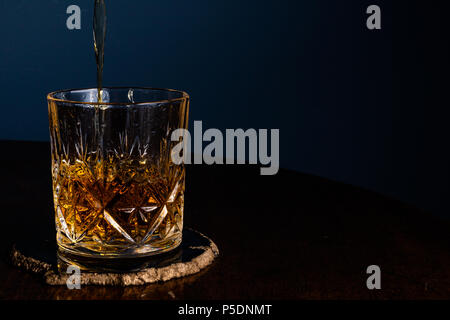 Whiskey in einem Kristall Tumbler auf einem dunklen Holztisch mit einem blauen Hintergrund gegossen. Stockfoto