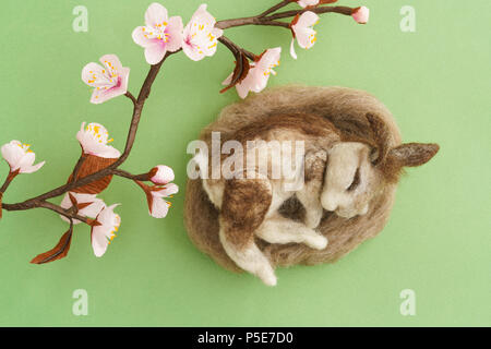 Nadel gefilzte Wilde Kaninchen schlafen in einem Nest auf grünem Hintergrund mit Krepppapier Pflaume Blüte Zweig Stockfoto