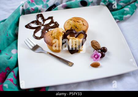 Muffin gefüllt mit Schokolade auf einem Teller, mit Schokolade Herz und Blume Zahlen-, Mandel-, Walnuß eingerichtet und violette Blume. Stockfoto