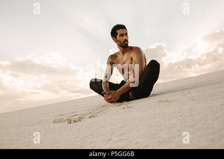 Athleten zu tun Stretching Übungen sitzen auf Sand Dune in Abend. Runner sitzt in der Wüste mit Füßen zusammen und Wegsehen. Stockfoto