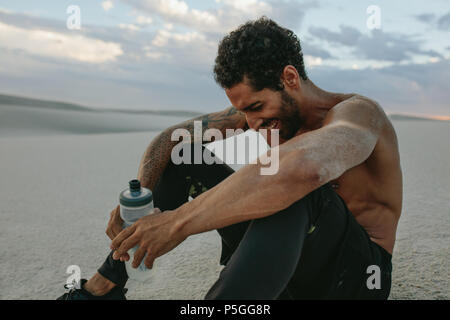 Happy männlichen Athleten sitzen auf Sand dune mit Wasserflasche und lächelnd in Abend. Mann Entspannung nach dem Training in der Wüste. Stockfoto