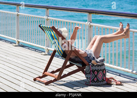 Bournemouth, Dorset, Großbritannien. Juni 2018. Wetter in Großbritannien: Sonnenanbeter begeben sich an einem weiteren schönen, sonnigen Tag mit ungebrochenem blauem Himmel und Sonnenschein zu den Stränden von Bournemouth. Eine schöne kühlende Brise macht die Hitze heute erträglicher. Frau, die sich entspannt und die Sonne genießt, sitzt auf dem Liegestuhl am Pier. Stockfoto