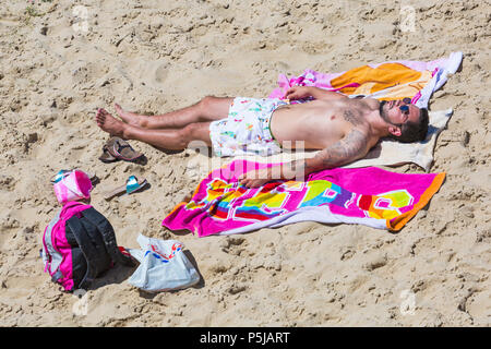 Bournemouth, Dorset, Großbritannien. Juni 2018. Wetter in Großbritannien: Sonnenanbeter begeben sich an einem weiteren schönen, sonnigen Tag mit ungebrochenem blauem Himmel und Sonnenschein zu den Stränden von Bournemouth. Eine schöne kühlende Brise macht die Hitze heute erträglicher. Mann trägt Shorts mit Papageien beim Sonnenbaden am Strand. Stockfoto