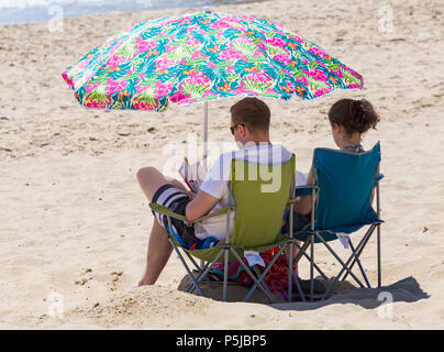 Bournemouth, Dorset, Großbritannien. Juni 2018. Wetter in Großbritannien: Sonnenanbeter begeben sich an einem weiteren schönen, sonnigen Tag mit ungebrochenem blauem Himmel und Sonnenschein zu den Stränden von Bournemouth. Eine schöne kühlende Brise macht die Hitze heute erträglicher. Junge Paare, die unter Sonnenschirm sitzen und Bücher lesen. Stockfoto