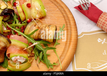 Gegrillte Zucchini mit Tomaten, Rucola, Schinken und Mozzarella Käse, mit Olivenöl und Balsamico Essig beträufelt. Zucchini auf runden Holzbrett. Stockfoto