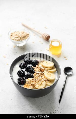 Frühstück smoothie Schüssel mit Müsli, Brombeeren, Bananen und Honig auf grauem Beton Hintergrund. Gesunder Lebensstil, Diät, vegetarische Kost Konzept Stockfoto