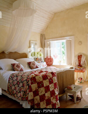 Land Schlafzimmer weiß voile Moskitonetz über dem antiken Bergere Bett mit rot+weiss Patchwork und Rose - gemusterte Steppdecken Stockfoto
