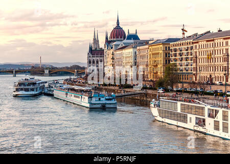 Kreuzfahrtschiffe bei Sonnenuntergang auf der Donau. Warm Sky auf dem Wasser widerspiegeln. Berühmte Parlament und Stadt für den Hintergrund. Budapest, Ungarn Stockfoto