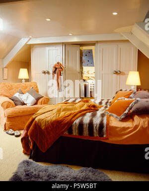 Orange Bettdecke und Bettwäsche auf dem Bett mit Fellimitat werfen auf Bett im Schlafzimmer im Dachgeschoss mit brennenden Lampen und orange Sessel Stockfoto