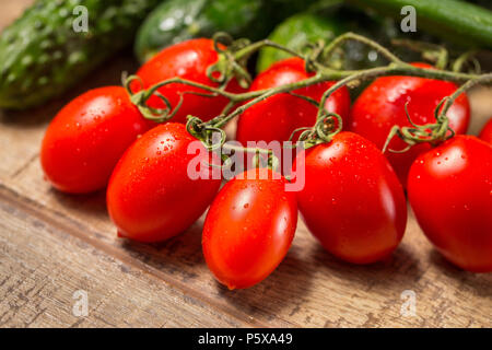 Gruppe von Pflaume Roma Tomaten auf einem Weinstock reif, auf einem Holztisch und Gurken im Hintergrund Stockfoto