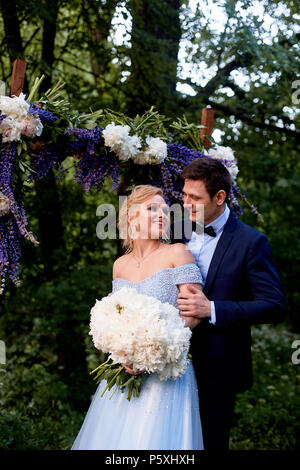 Die Braut und der Bräutigam stand auf der Arch, mit Blumen geschmückt, mit einem großen Blumenstrauß aus den weißen Pfingstrosen. Registrierung vor Ort im Park Hochzeit gehen ein Stockfoto