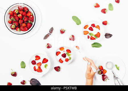 Die weibliche Hand, die Löffel über einer Schale Erdbeereis unter roten Beeren, Minze und Basilikum, Teller mit Desserts, Ansicht von oben. Flach kom Stockfoto