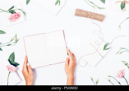 Woman's Hände halten ein aufgeschlagenes Buch unter weiblichen Schreibtisch mit blühenden Blumen, grünen Blättern und weißen Geschenkboxen, mit Garn gebunden, Ansicht von oben. Festliche Flo Stockfoto