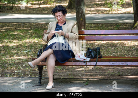 Niska Banja, Serbien - 25. Juni, 2018: ältere Dame sitzt auf der Bank im Park und stricken Tischdecke. Alte Frau häkeln mit weißem Garn und Nadeln Stockfoto