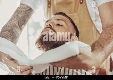 Friseur deckt das Gesicht eines Mannes mit Bart mit einem warmen Handtuch. Foto im Vintage Style Stockfoto