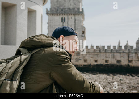 Porträt eines jungen Touristen Kerl oder Reisenden Mann mit einem Rucksack, der sitzt und starrt in die Ferne in Lissabon in Portugal. Belem Turm im Hintergrund Stockfoto