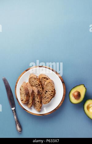 Gehackte frische französische Baguette auf den Teller, Messer und Avocado auf blauem Hintergrund. Ansicht von oben, kopieren. Stockfoto